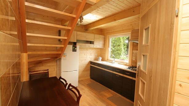 A wooden cabin interior with a dining table, chairs, a kitchen area with a window, and a wooden staircase. - Agroturystyka Katarzyna Tarasiewicz (Suwalki)