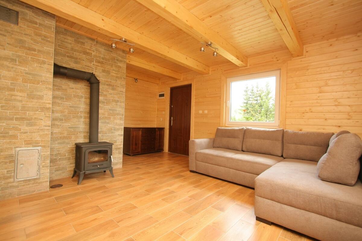 A log cabin interior with a stone fireplace, wooden flooring, a sofa, and a window with a view of trees.