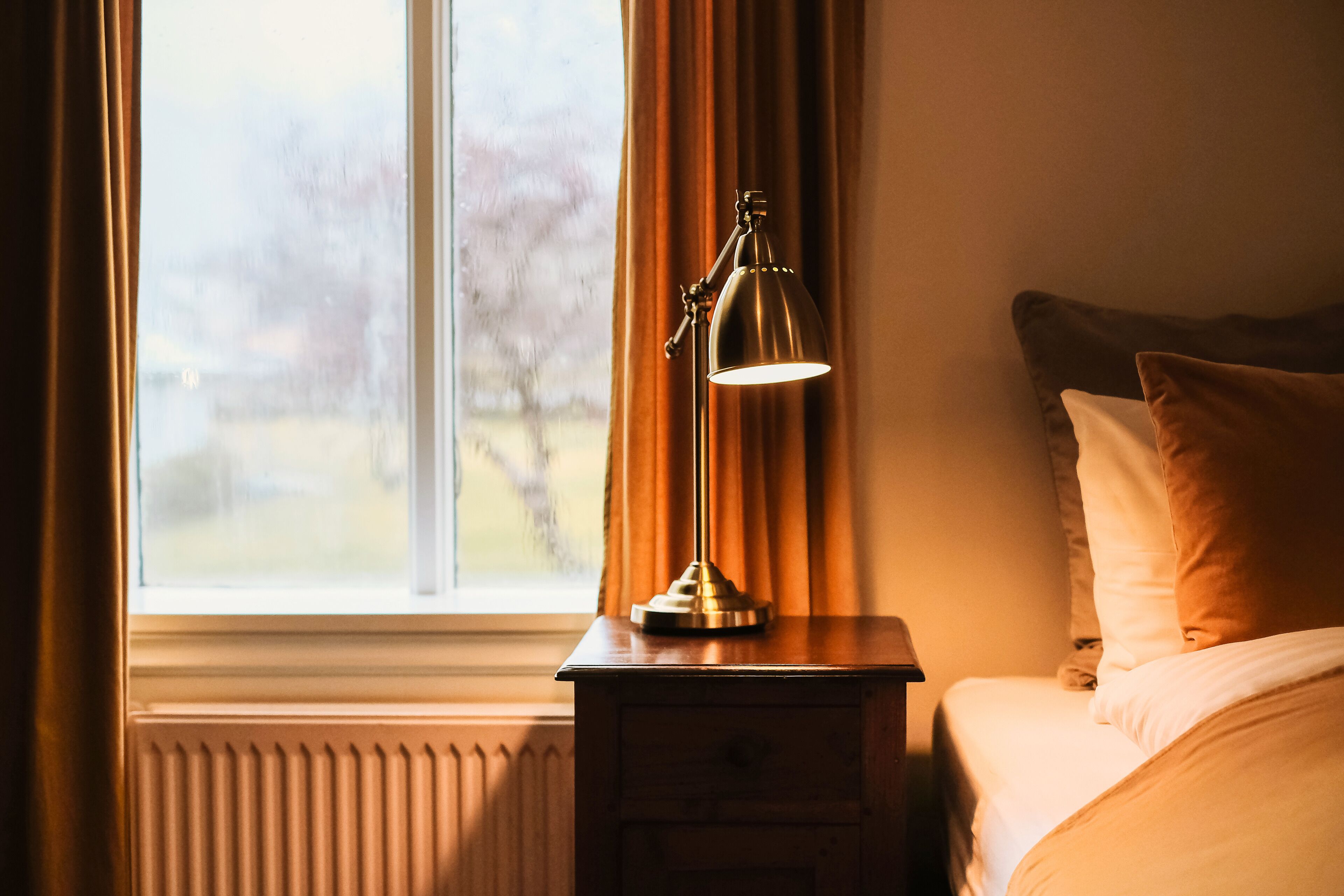 Ein Zimmer mit Nachttisch, Lampe, einem Fenster mit Vorhängen und einem Bett mit Kissen.