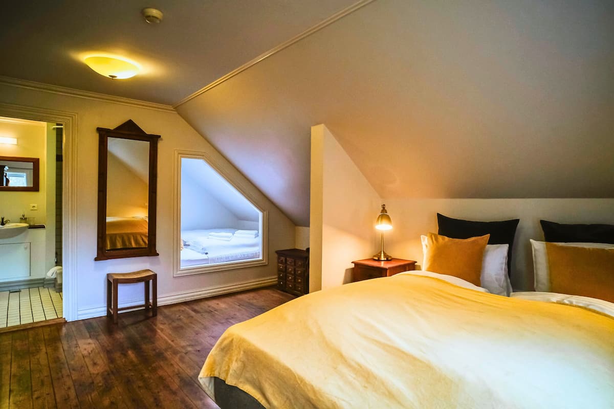 A bedroom with a large bed, a nightstand with a lamp, a window with a view, a mirror, and a small stool.