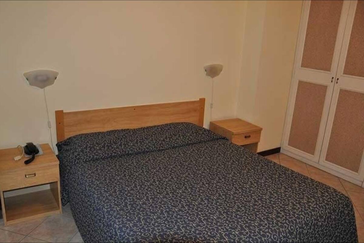 A bedroom with a wooden bed, a bedside table with a lamp, a wardrobe, and a telephone on the nightstand.