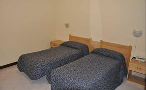 Una habitación pequeña y sencilla con dos camas individuales, cabeceros de madera, mesitas de noche y una pared lisa.