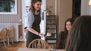 Una persona sirve un postre en una mesa dentro de un restaurante, con un menú en una pizarra en la pared.