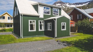Una casa de dos pisos con fachada verde y detalles blancos, que presenta un ventanal destacado y un techo inclinado.