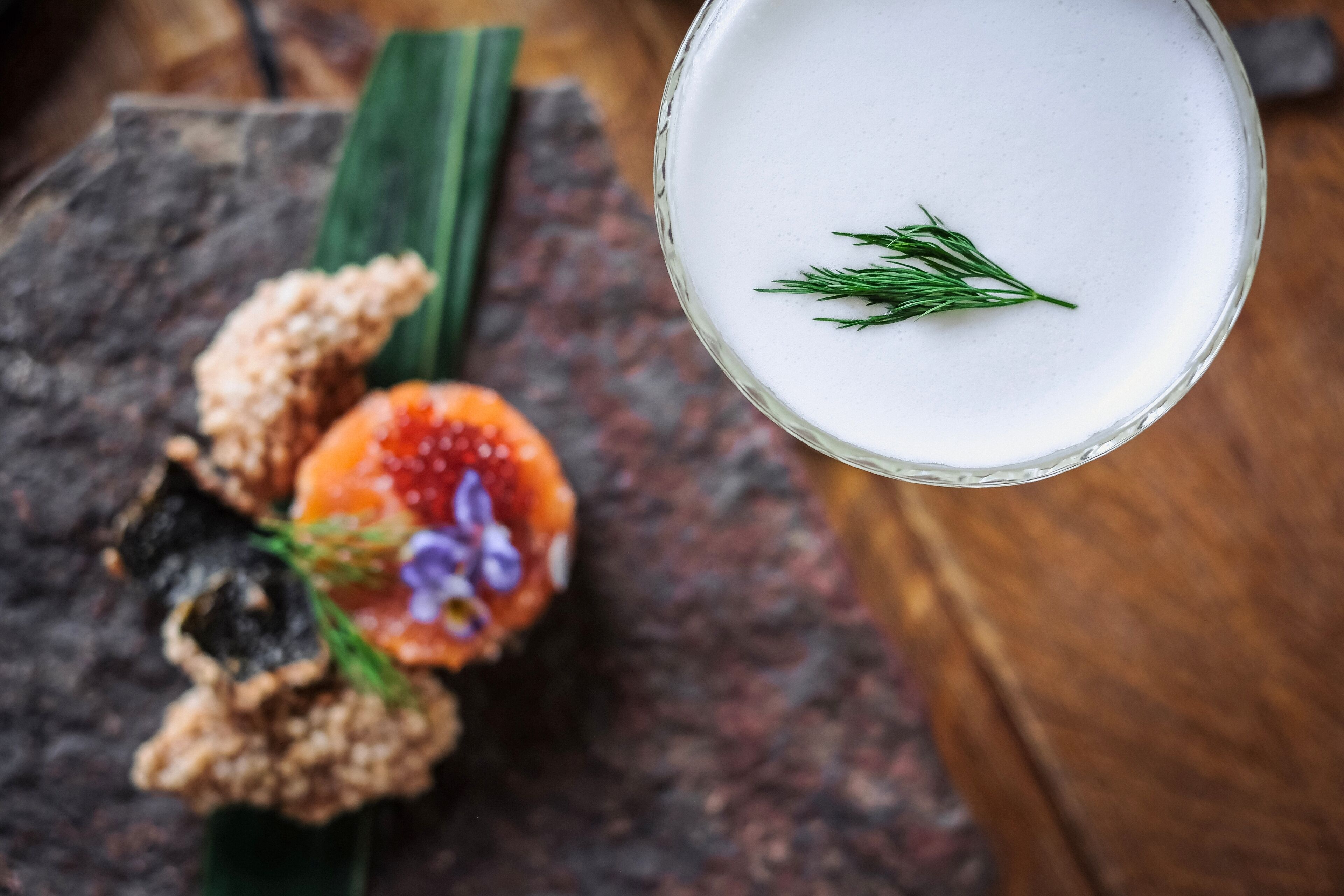 A glass of white beverage garnished with a sprig of dill, placed on a dark stone surface with a decorative food item beside it.