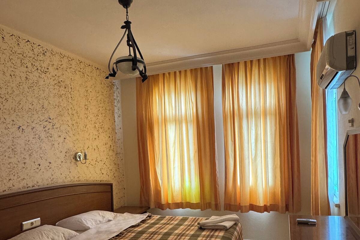 Hotellihuone, jossa on sänky, yöpöytä, lamppu, verhot ja ikkuna.