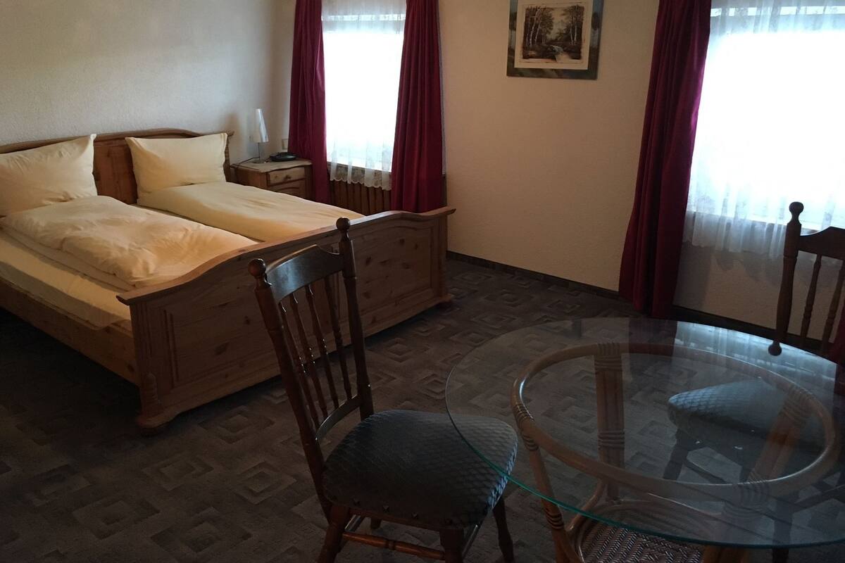 Een hotelkamer met een tweepersoonsbed, een glazen tafel, twee stoelen, een nachtkastje, een lamp en een schilderij aan de muur.