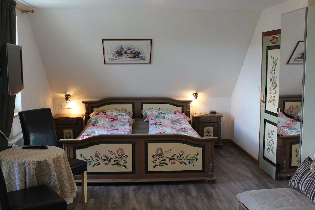Een slaapkamer met een houten bed, twee nachtkastjes met lampen, een stoel, een klein tafeltje met tafelkleed en een tv aan de muur.