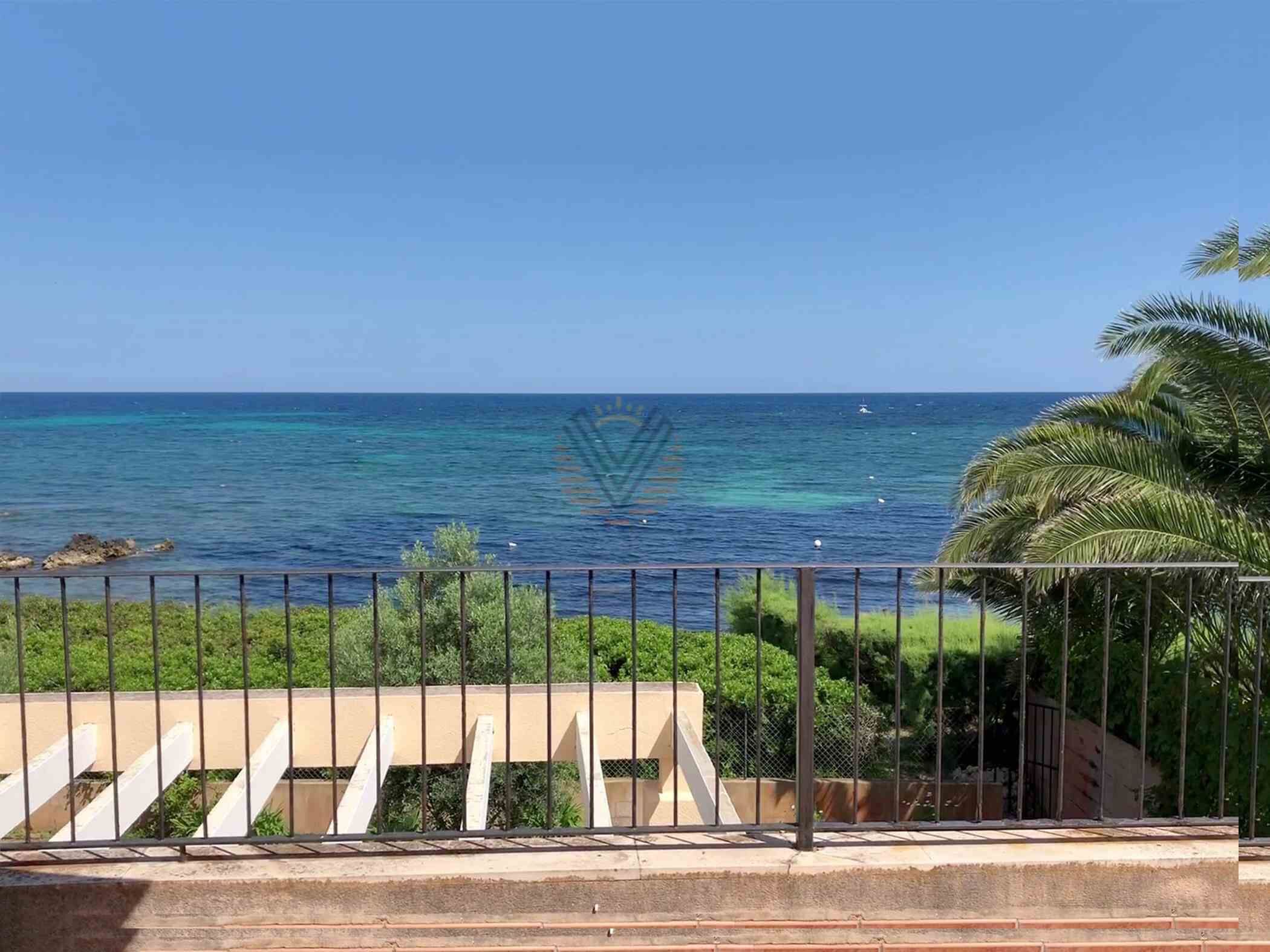 A coastal view with a clear blue sea, a balcony with a metal railing, and greenery.