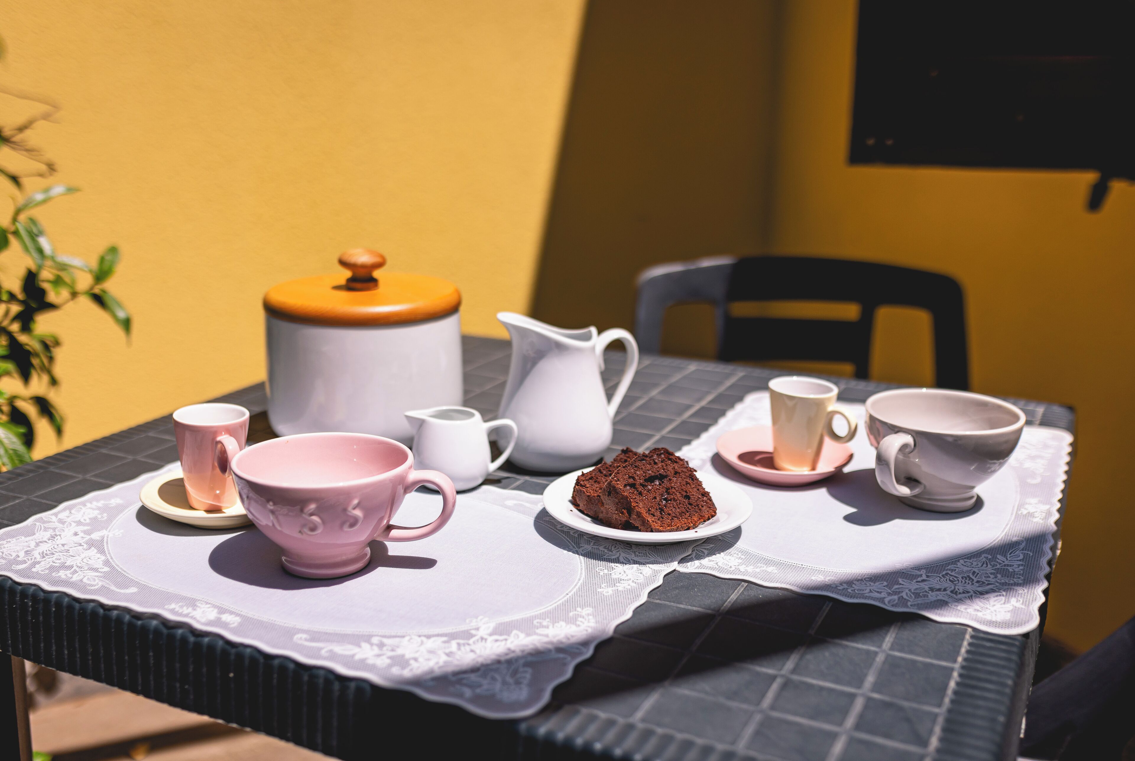 Een tafel met een kantkleed, een witte karaf, een roze mok, een roze kom, een bord met een plak cake en een witte theepot met een geel deksel.