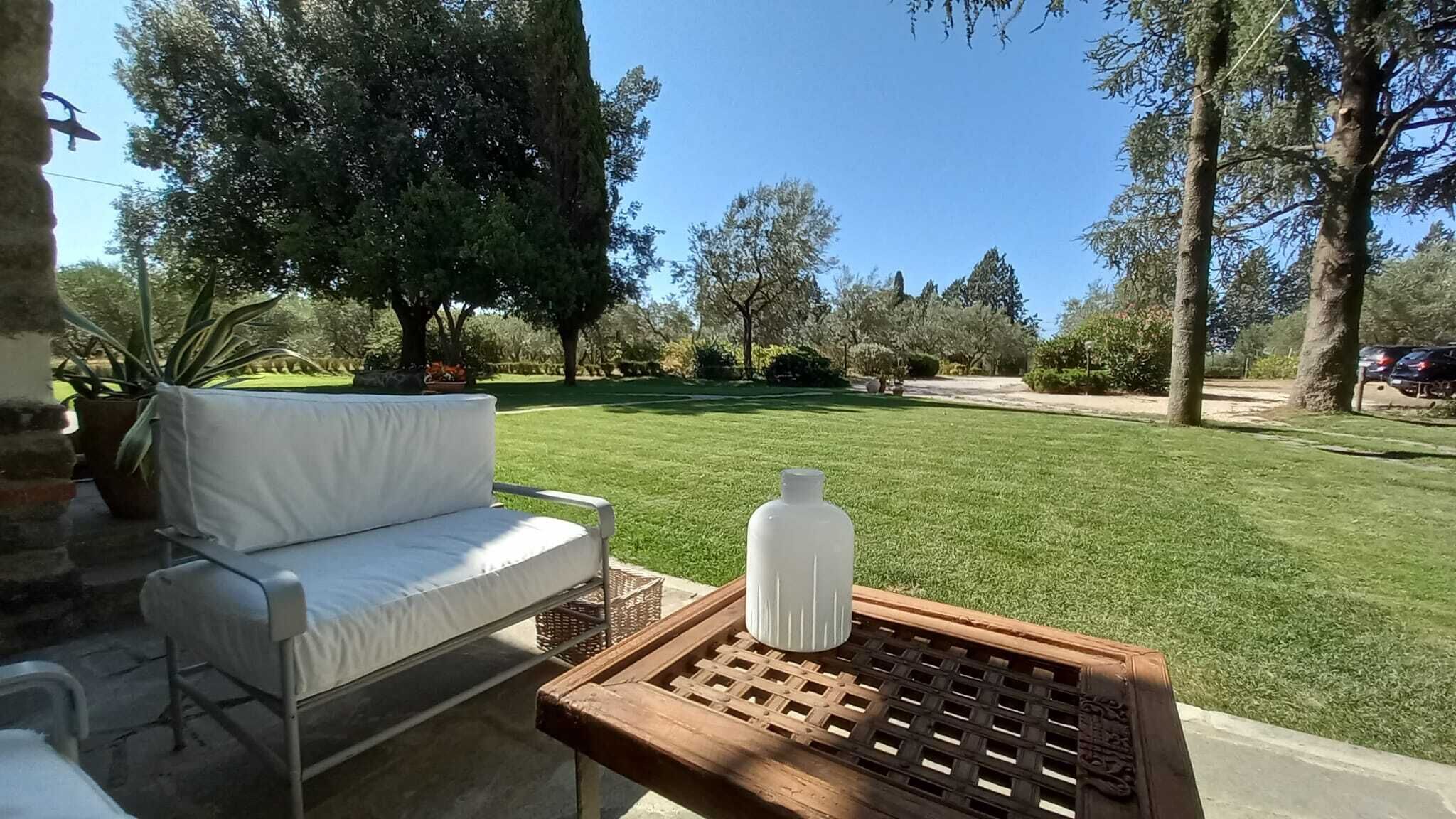A patio area with a white cushioned chair, a wooden table with a bottle, and a well-maintained lawn.