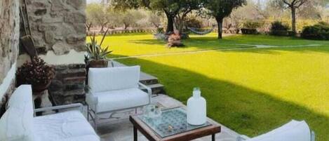 Un patio con mobiliario blanco, un muro de piedra y vistas a un césped y árboles bien cuidados.