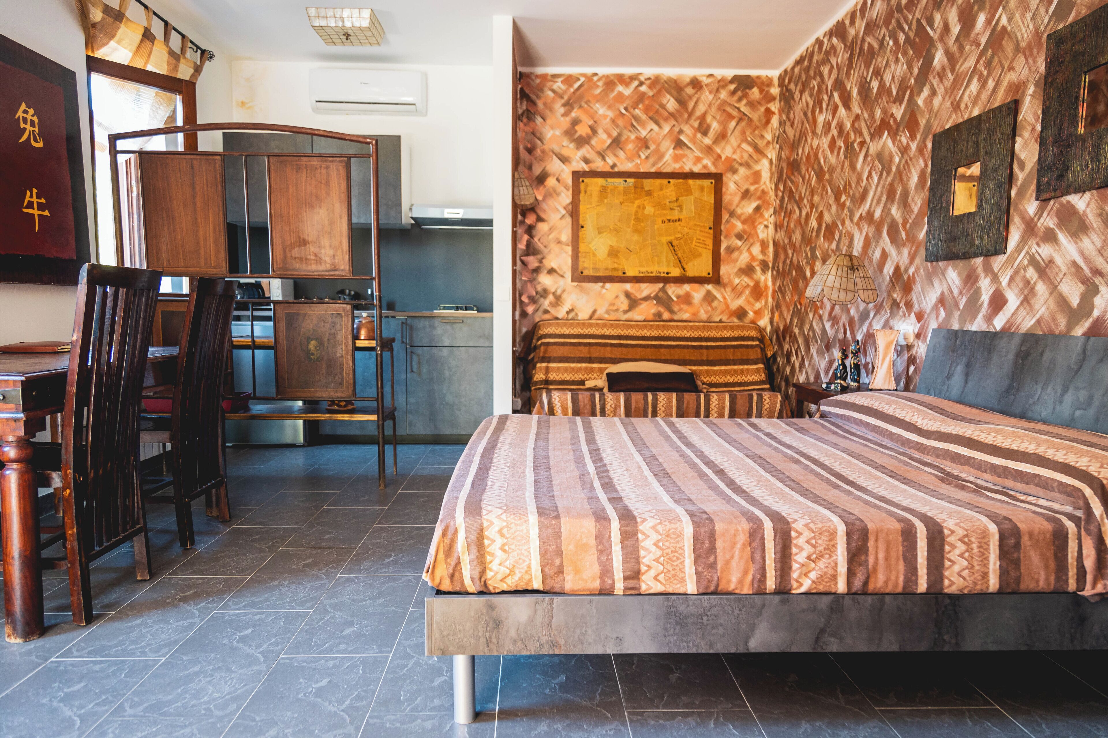 A hotel room with a bed, a dining table with chairs, a kitchen area, and a patterned wall.