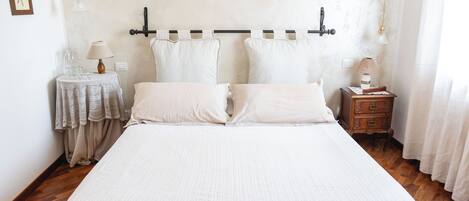A neatly made bed with a white bedspread and pillows, a wooden nightstand with a lamp, and a small table with a lamp and a pitcher.