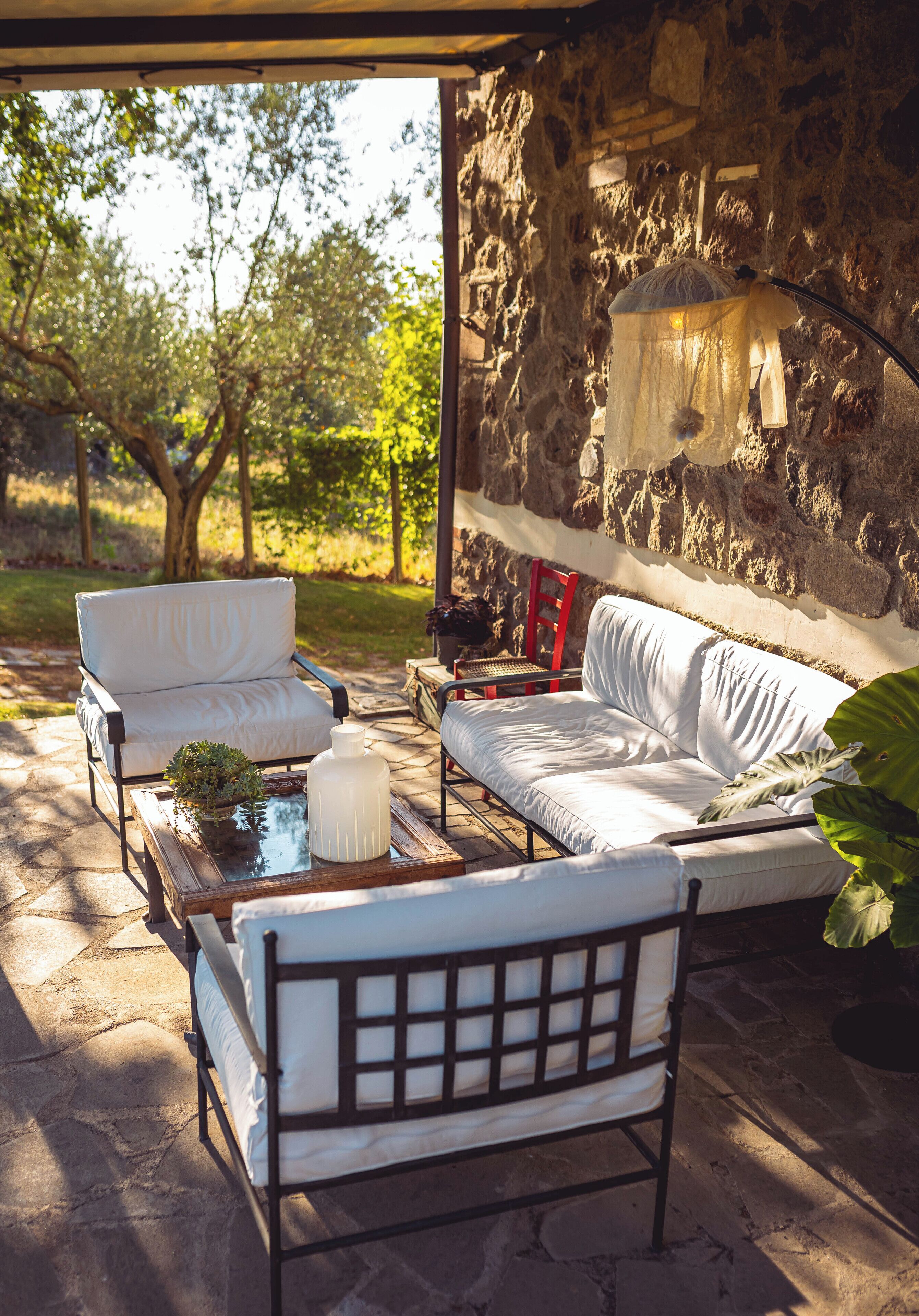 A covered outdoor seating area with stone walls, a white sofa, a chair, and a table with a plant.