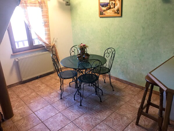 A dining area with a round table and four chairs, a radiator, a window with curtains, and a painting on the wall.