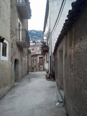 Un estrecho callejón entre edificios con paredes de piedra, algunos balcones y una montaña al fondo.