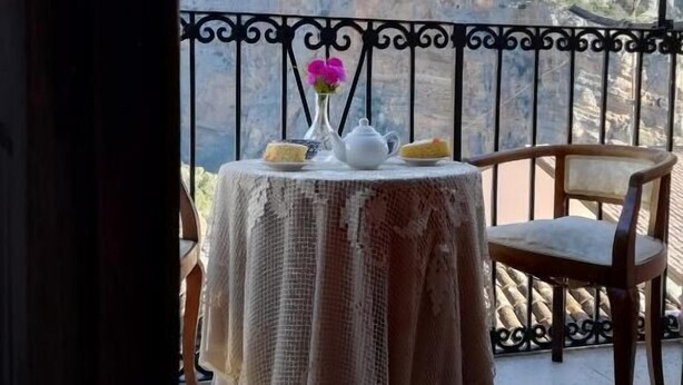 Une table recouverte d’une nappe en dentelle, un vase rempli de fleurs, une théière et une chaise sur un balcon offrant une vue sur la montagne.