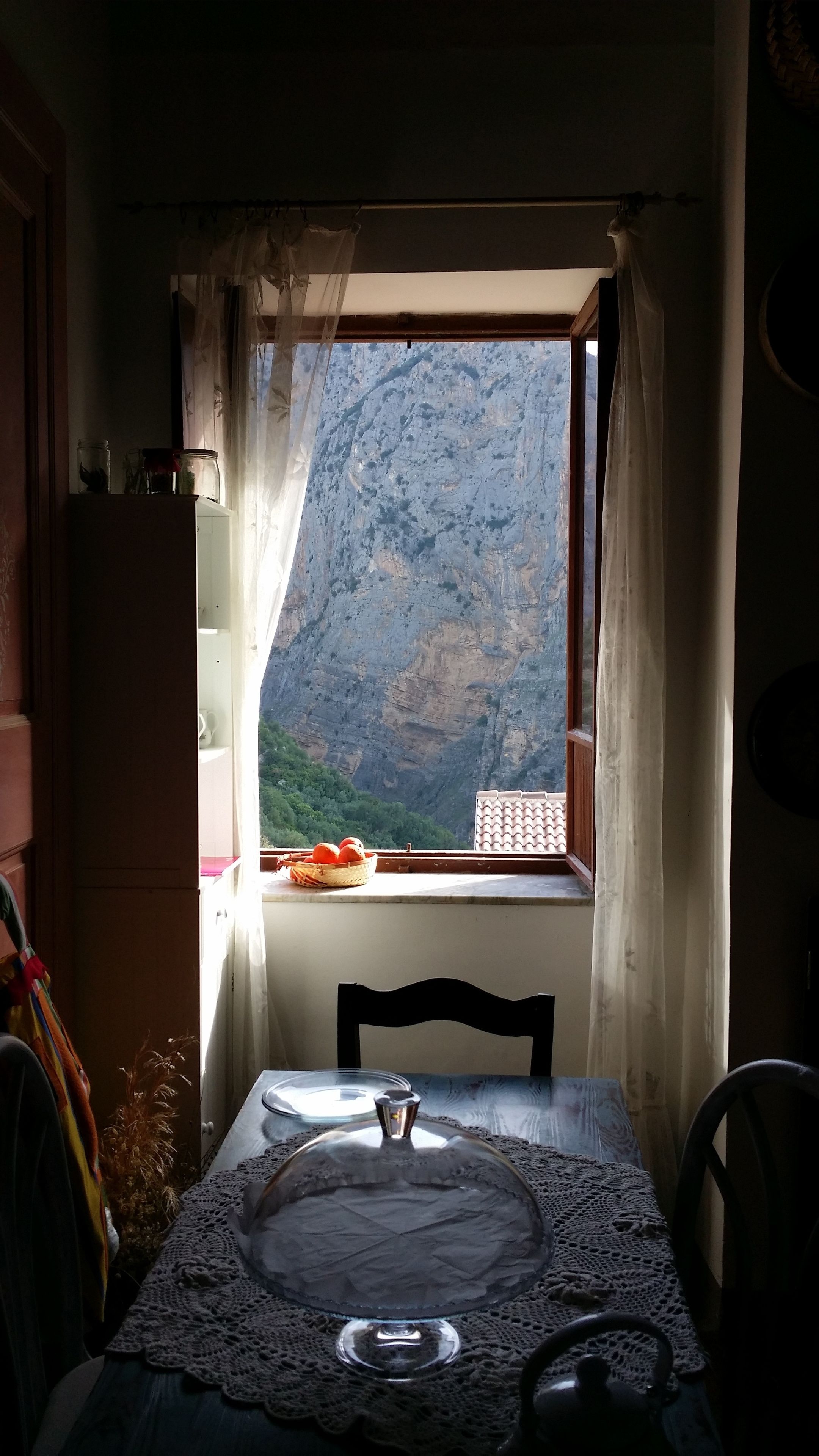A dining area with a table set for two, a view of a mountainous landscape through a window, and a vase with dried plants on the table.
