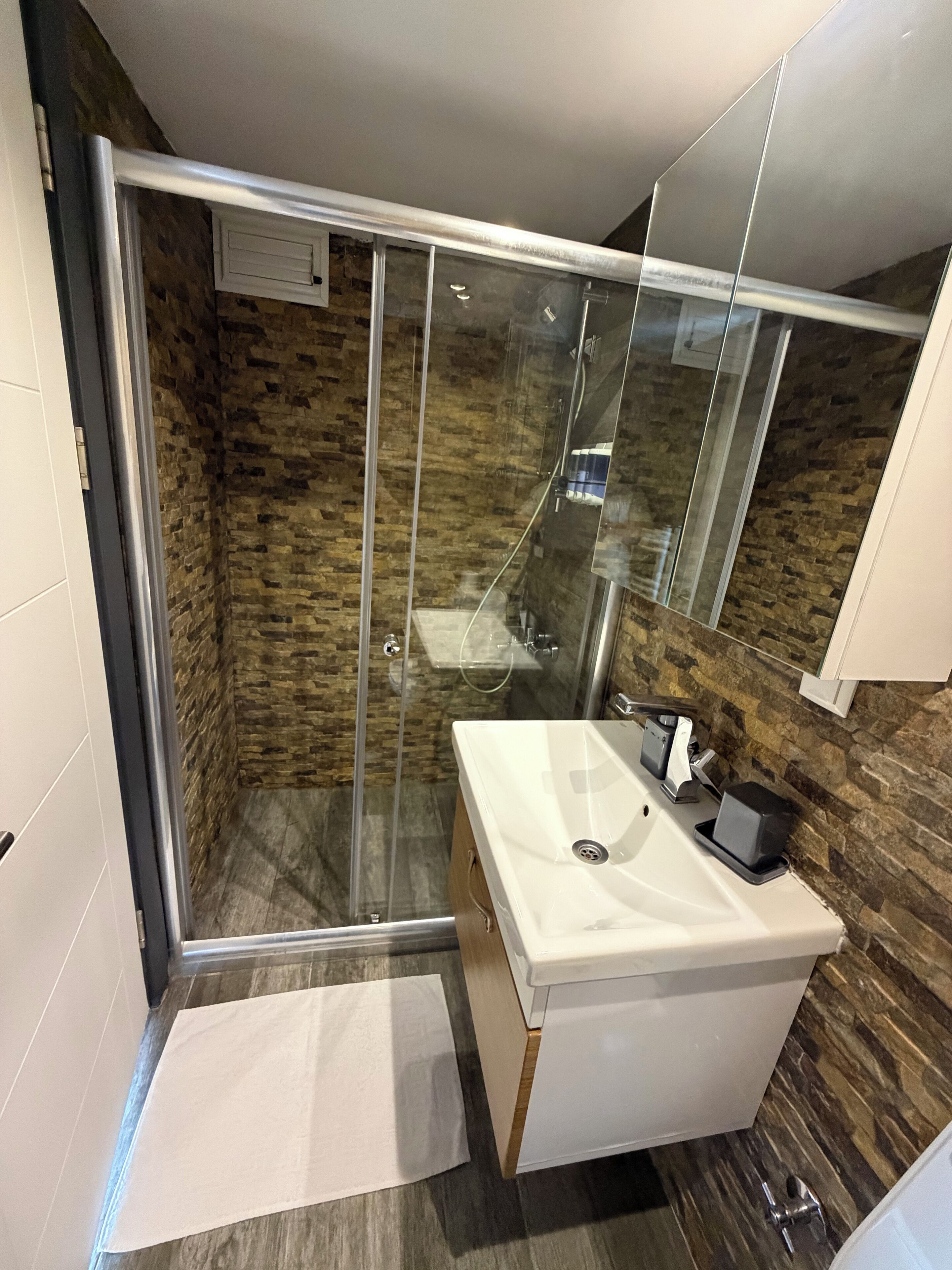 Een moderne badkamer met een douchecabine van glas, een witte wastafel en een houten opbergmeubel.