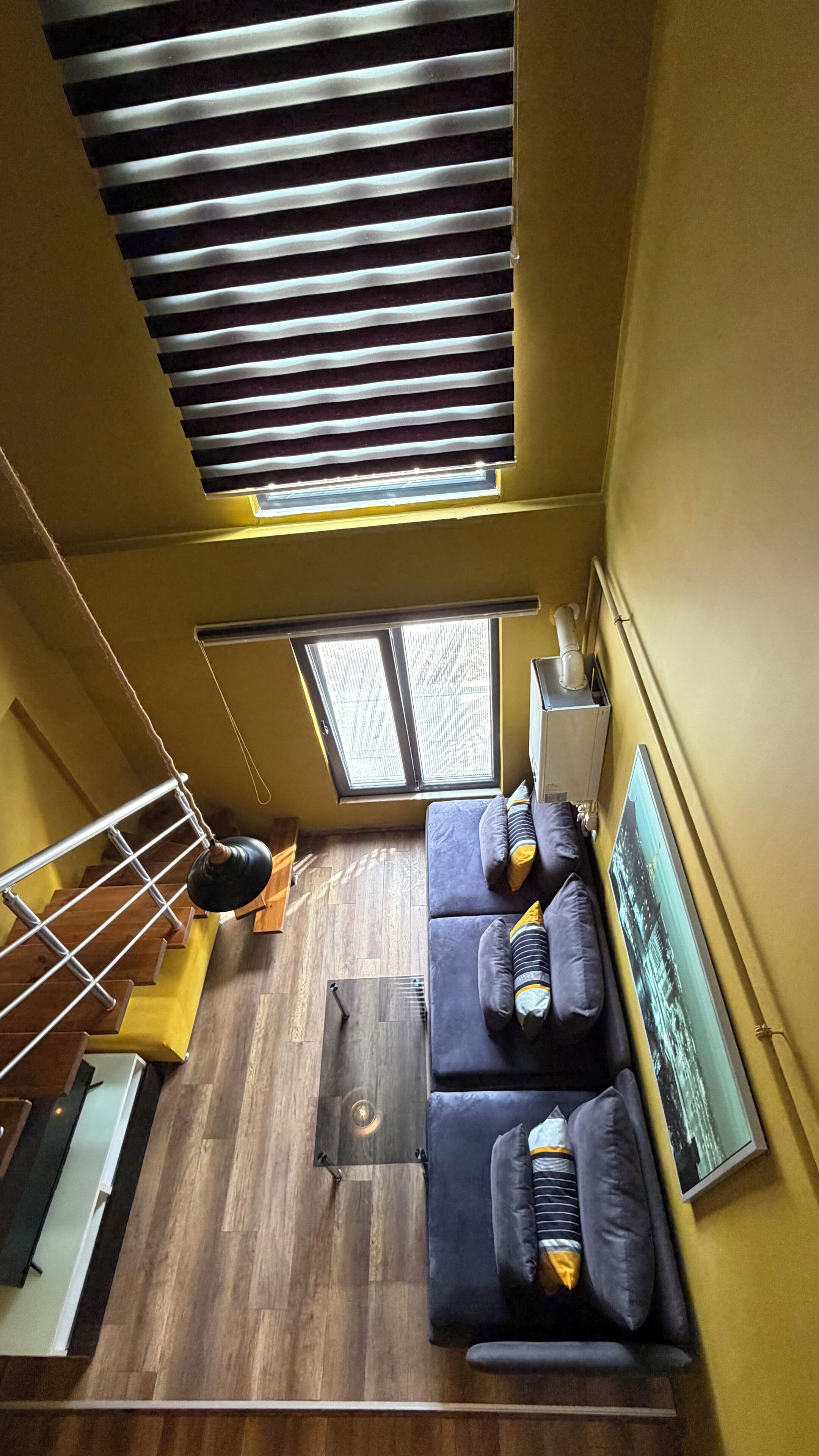 Una habitación con techos altos, suelo de madera, un sofá oscuro, una mesita y un ventanal con persianas.