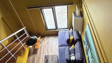 Una habitación con techos altos, suelo de madera, un sofá oscuro, una mesita y un ventanal con persianas.