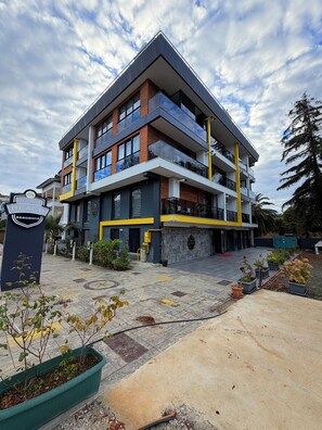 A modern multi-story hotel with a prominent entrance, outdoor seating area, and a landscaped garden. - Deluxe Beach Residence (Atakum)