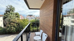 A balcony with two white chairs and a table, a wooden wall, and a view of buildings and trees. - Deluxe Beach Residence (Atakum)