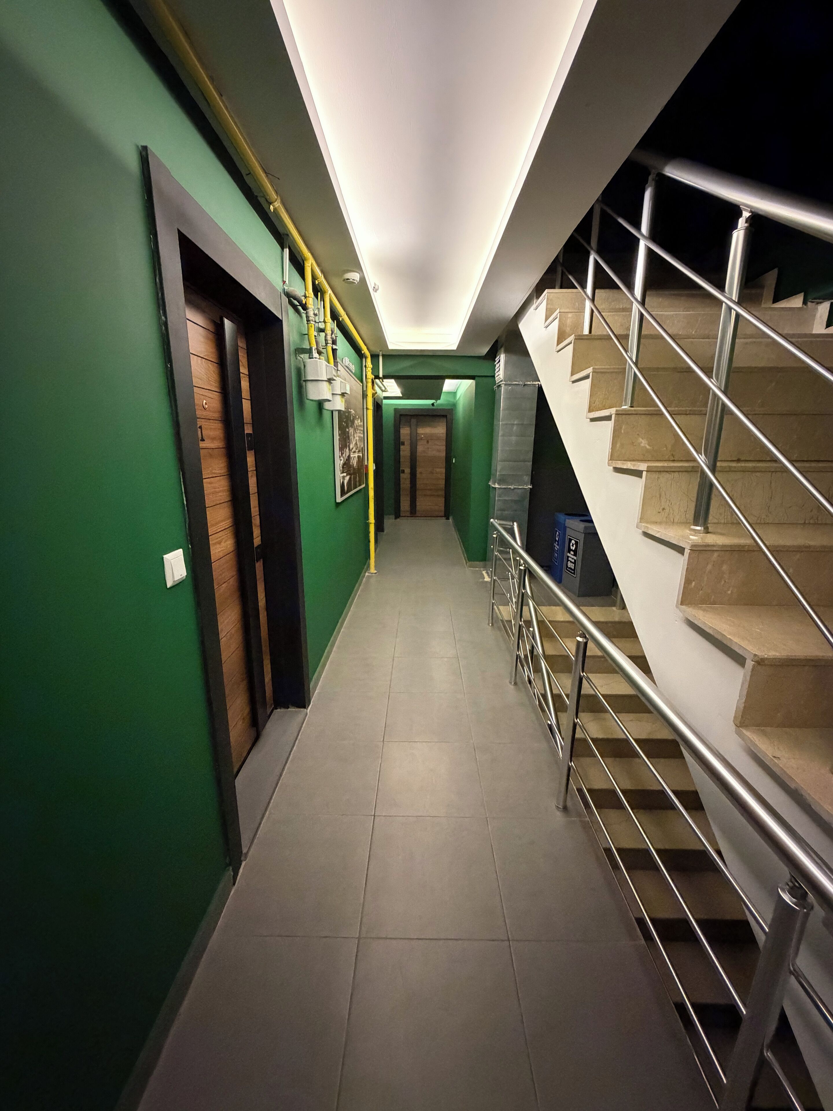 En hall med gröna väggar, en trädörr, en trappa med metallräcken och ett klinkergolv.