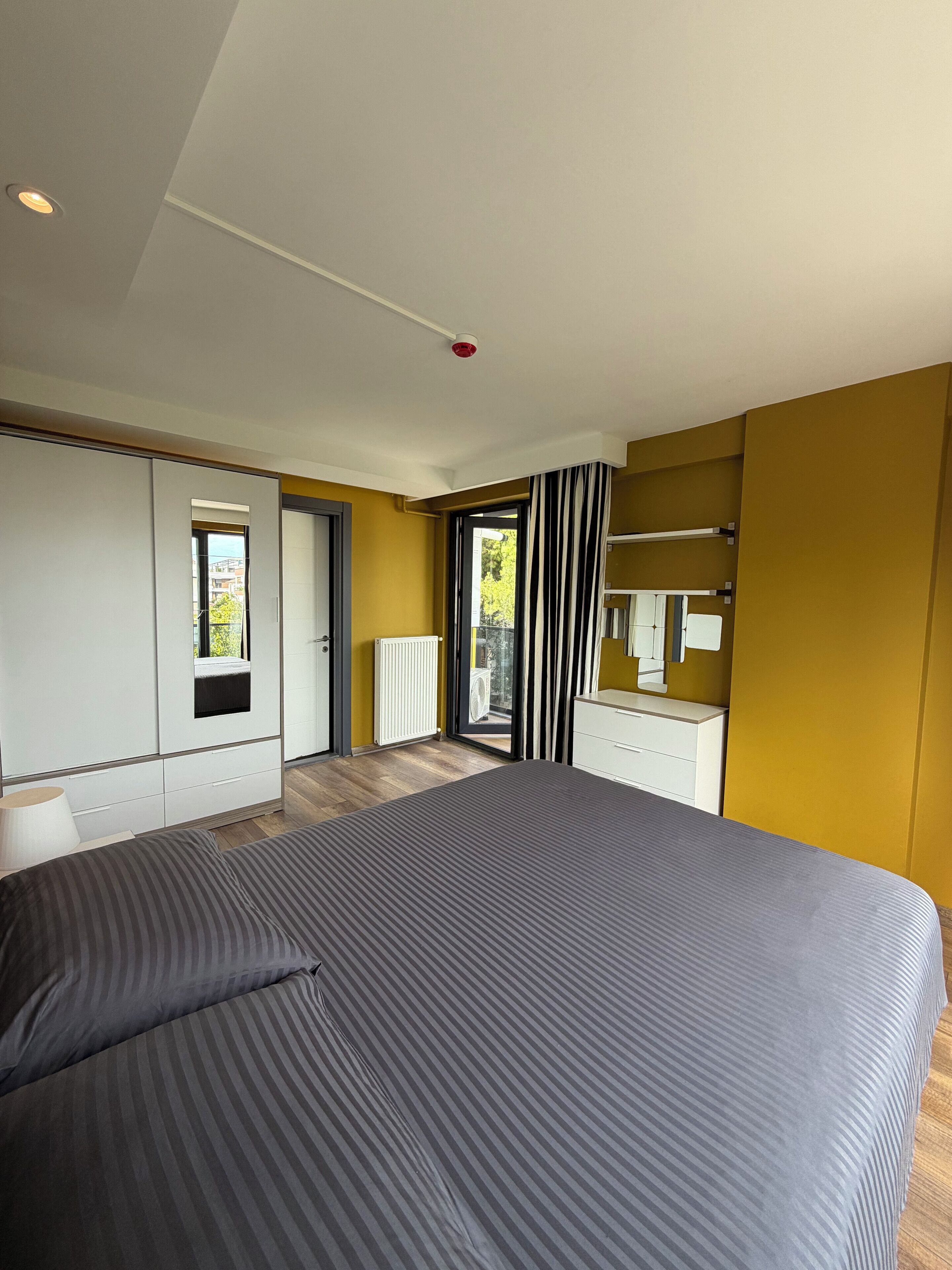 A modern bedroom with a large bed, a wardrobe, a mirror, and a view of the outdoors.