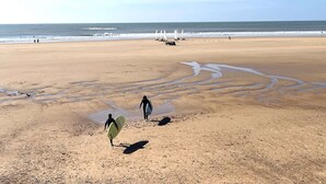 Une vaste plage de sable fin, avec des vagues douces, un ciel dégagé et quelques personnes qui se promènent.
