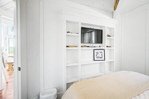 A bedroom with a TV mounted on a white wall, a bed with a beige blanket, and a door leading to a wooden-floored hallway. - Lighthouse - Oceanfront, stunning, updated classic (Cloverdale)