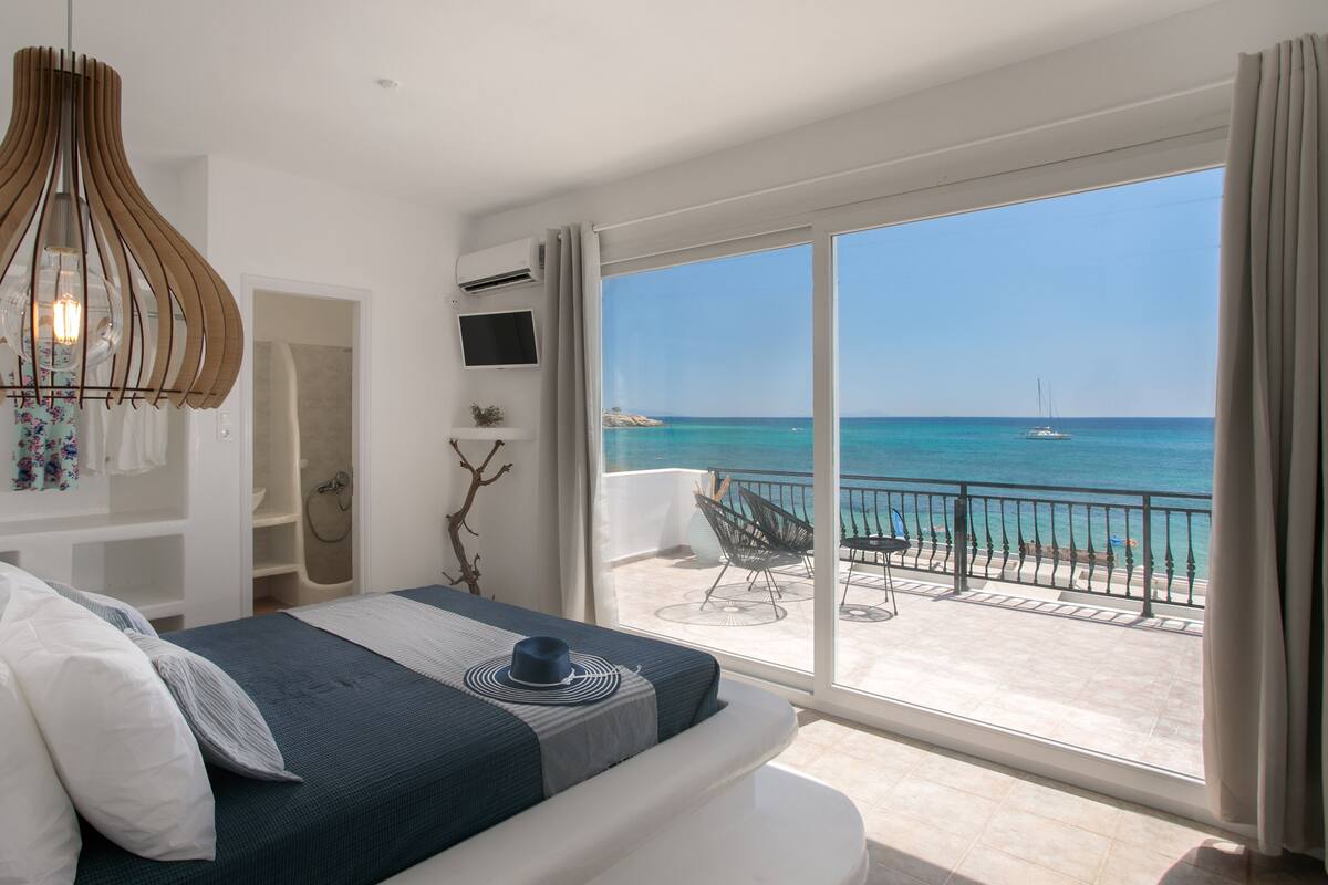 A bedroom with a large balcony, a bed, a TV, and a view of the sea.