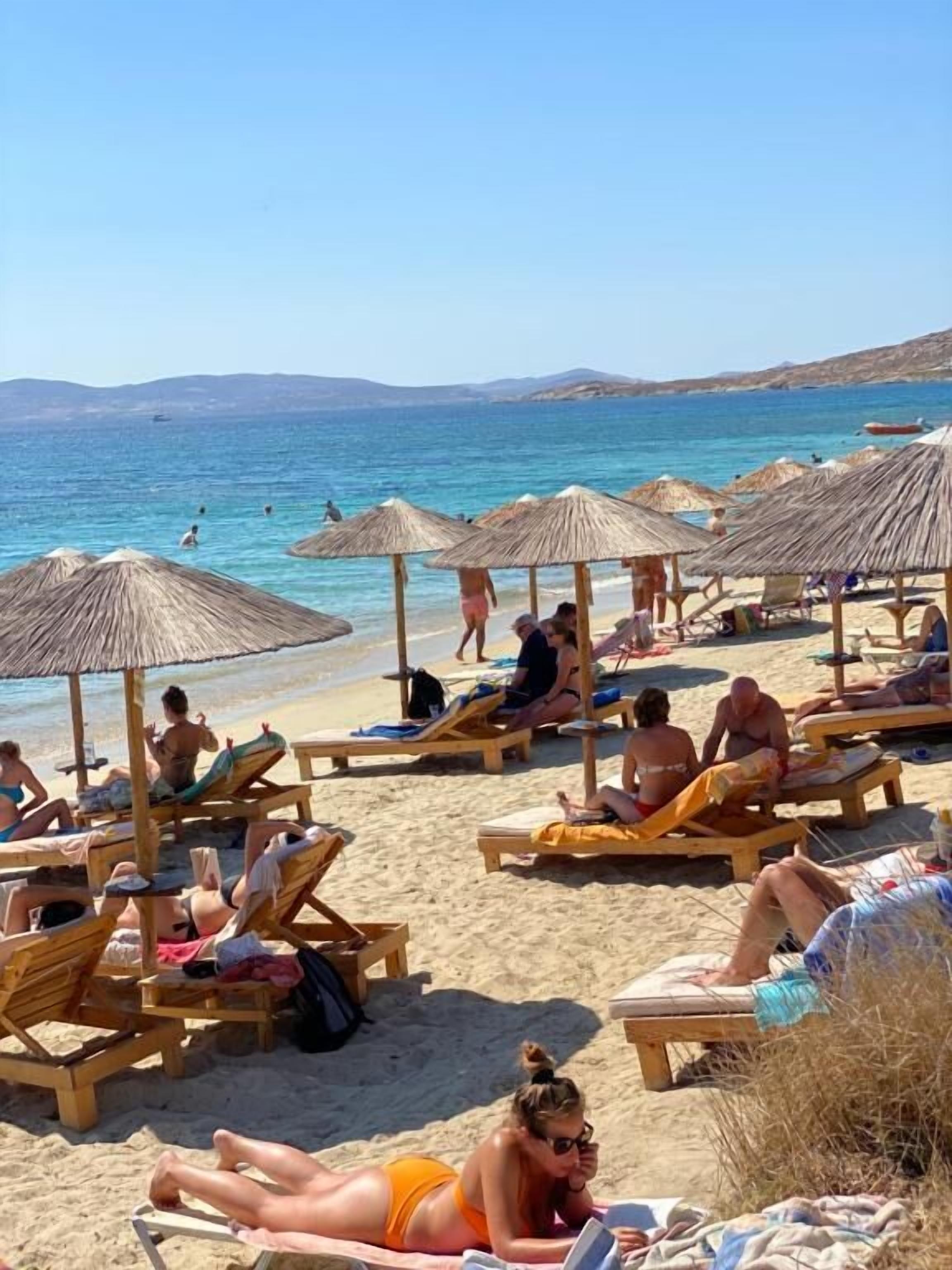 Une plage avec des transats et des parasols, des gens qui prennent un bain de soleil, et une vue dégagée sur la mer et les collines lointaines.