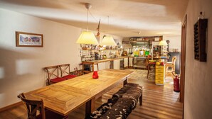 Un'ampia sala da pranzo con un tavolo di legno, panche e sedie. Si intravede in secondo piano una cucina con diversi elettrodomestici e utensili.