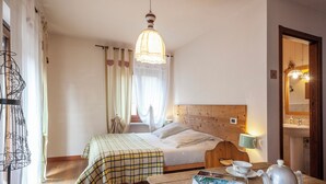 Un'accogliente camera da letto con una testiera in legno, un letto con coperta a quadri, un comodino con teiera e una rivista, e una finestra con tende leggere.