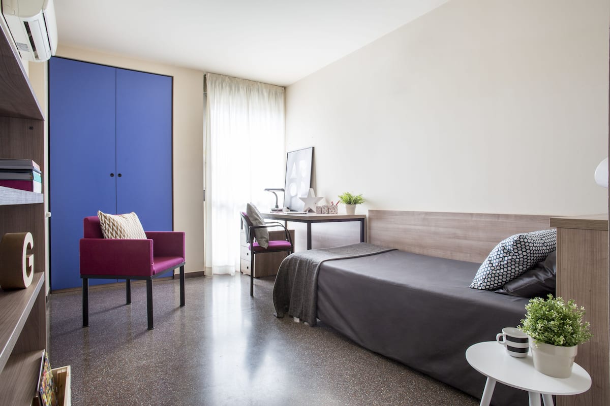 A compact hotel room with a bed, a desk, a chair, a wardrobe, and a small table with a plant.