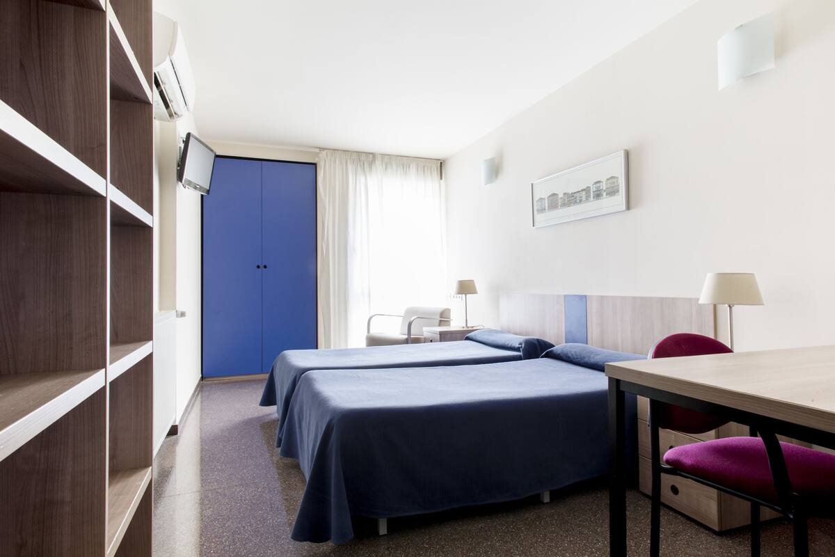 A hotel room with two beds, a desk, a chair, a lamp, and a TV.