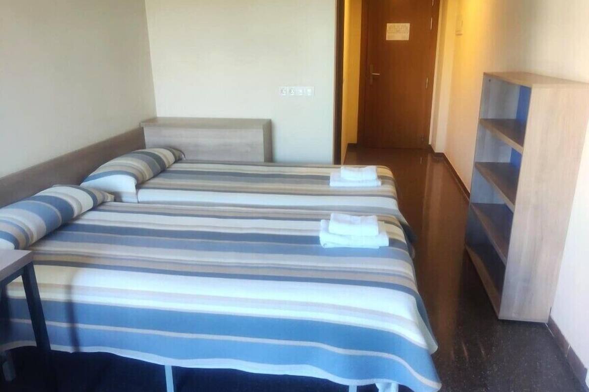 A hotel room with two beds, a wooden wardrobe, and a small shelf unit.