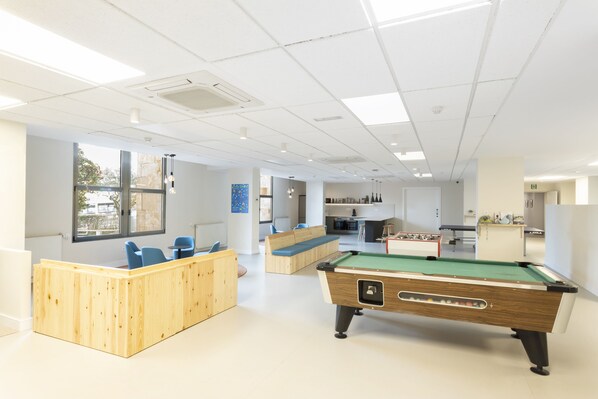 A modern indoor space with a pool table, seating areas, and a kitchen in the background. - Residencia Universitaria Resa Hernán Cortés - Centro Adscrito a REAJ (Salamanca)