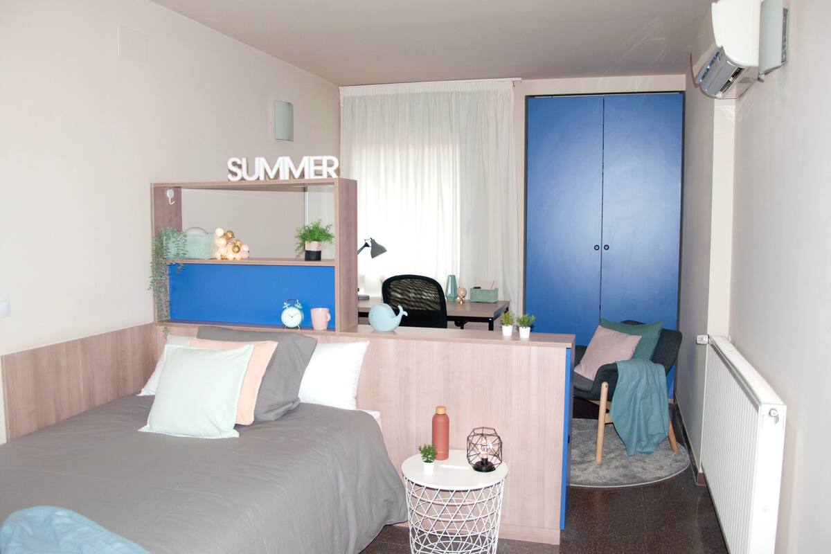 A bedroom with a bed, a desk, a chair, a small table, and a wardrobe.