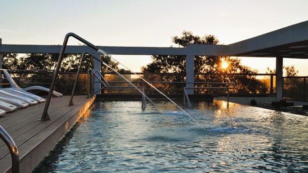 Una piscina con muelle de madera y barandillas de metal, rodeada de árboles y un atardecer al fondo.