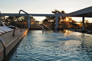 Une piscine dotée d’une terrasse en bois et de rampes en métal, entourée d’arbres, avec un coucher de soleil en arrière-plan.