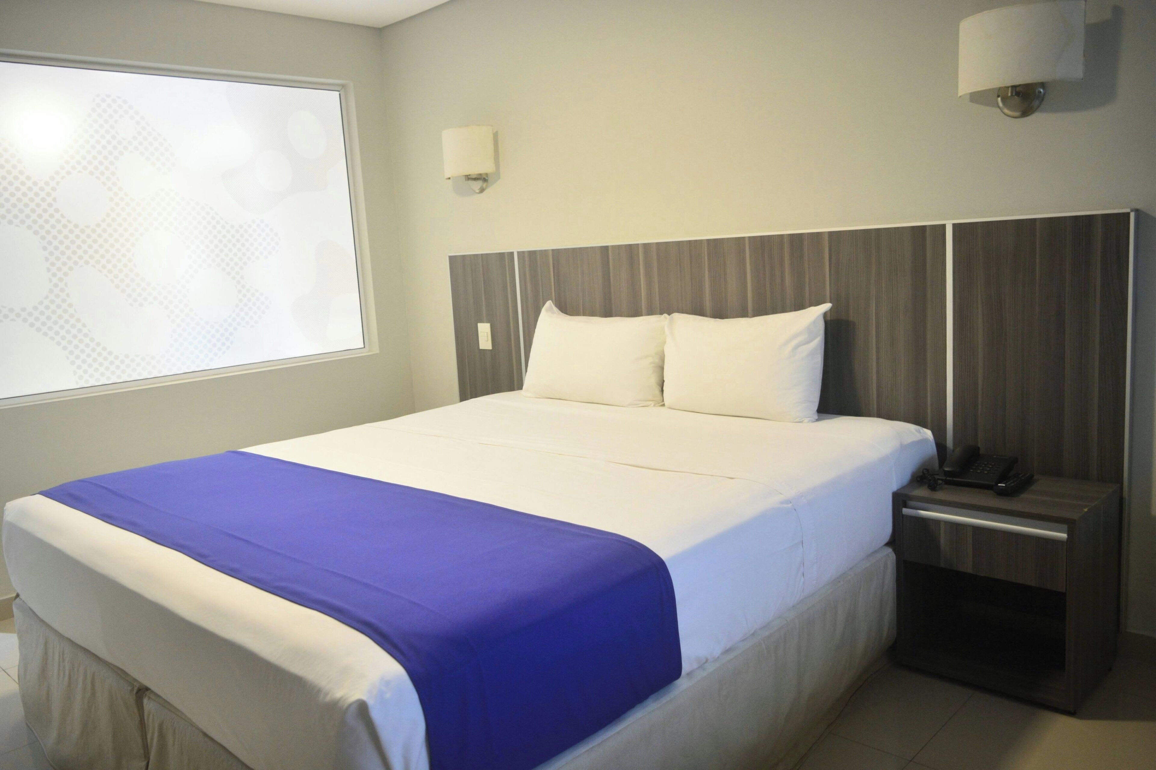 A hotel room with a large bed, a nightstand with a phone, and a window with blinds.