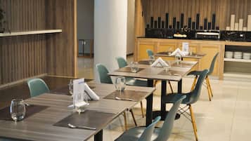 A dining area with multiple tables and chairs, a kitchen in the background, and a wooden wall.