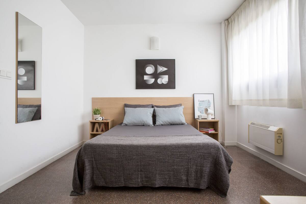 Moderni hotellihuone, jossa on suuri sänky, pääty, yöpöytä, seinään kiinnitetty lamppu ja ikkuna, jossa on verhot.