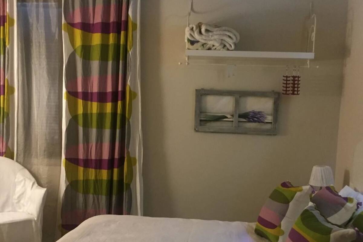 A bedroom with a bed, colorful striped pillows, a bedside table, a lamp, and a curtain with a checkered pattern.