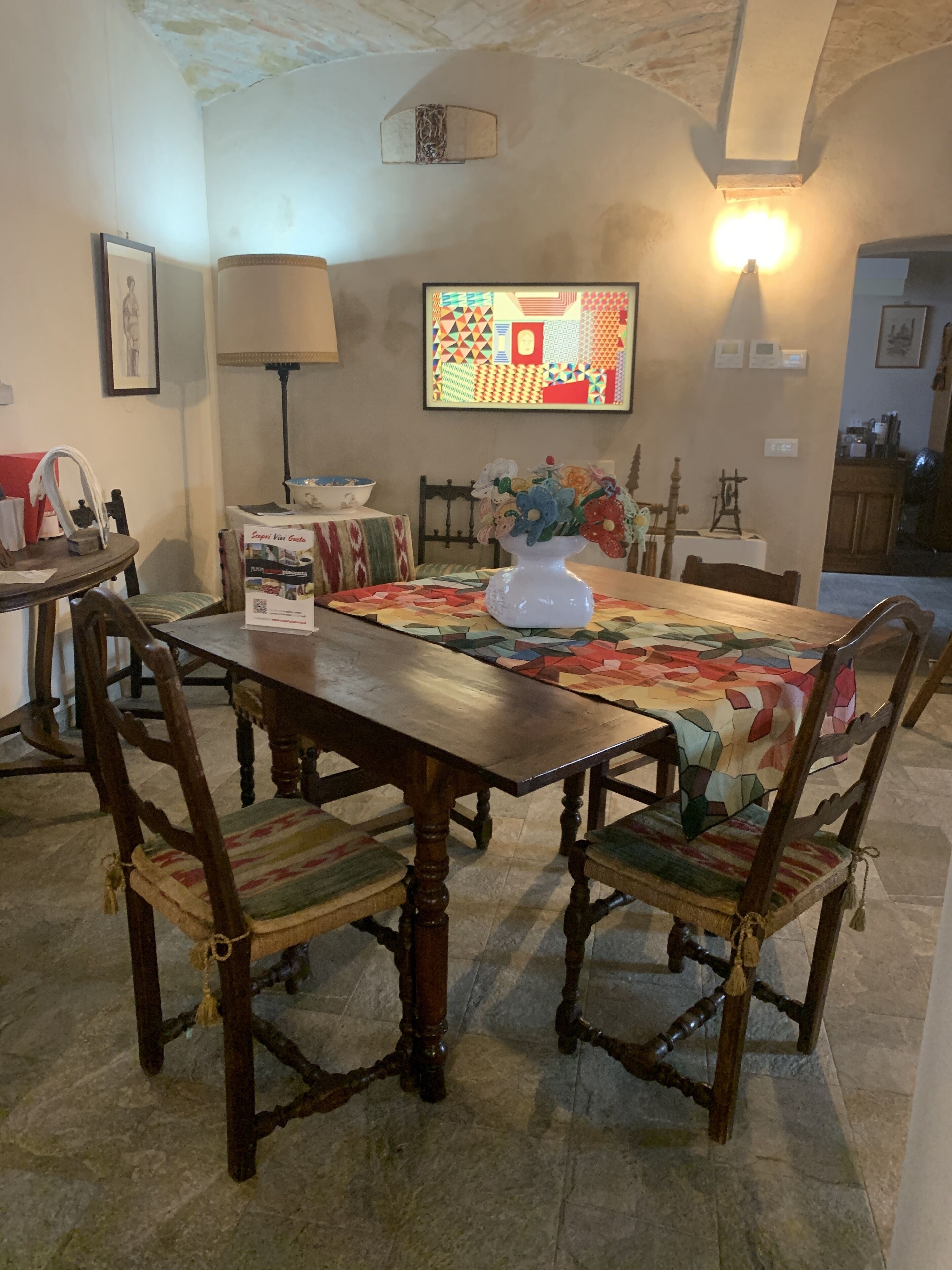 Un coin repas avec une table en bois, des chaises et un chemin de table à motifs. On aperçoit un vase rempli de fleurs sur la table, et une lampe à l’arrière-plan.