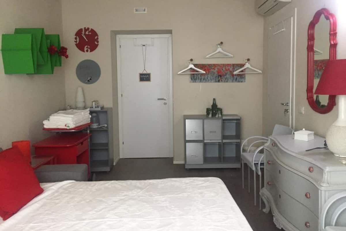 A hotel room with a bed, a desk with a chair, a dresser, a wardrobe, a mirror, a clock, and a door.