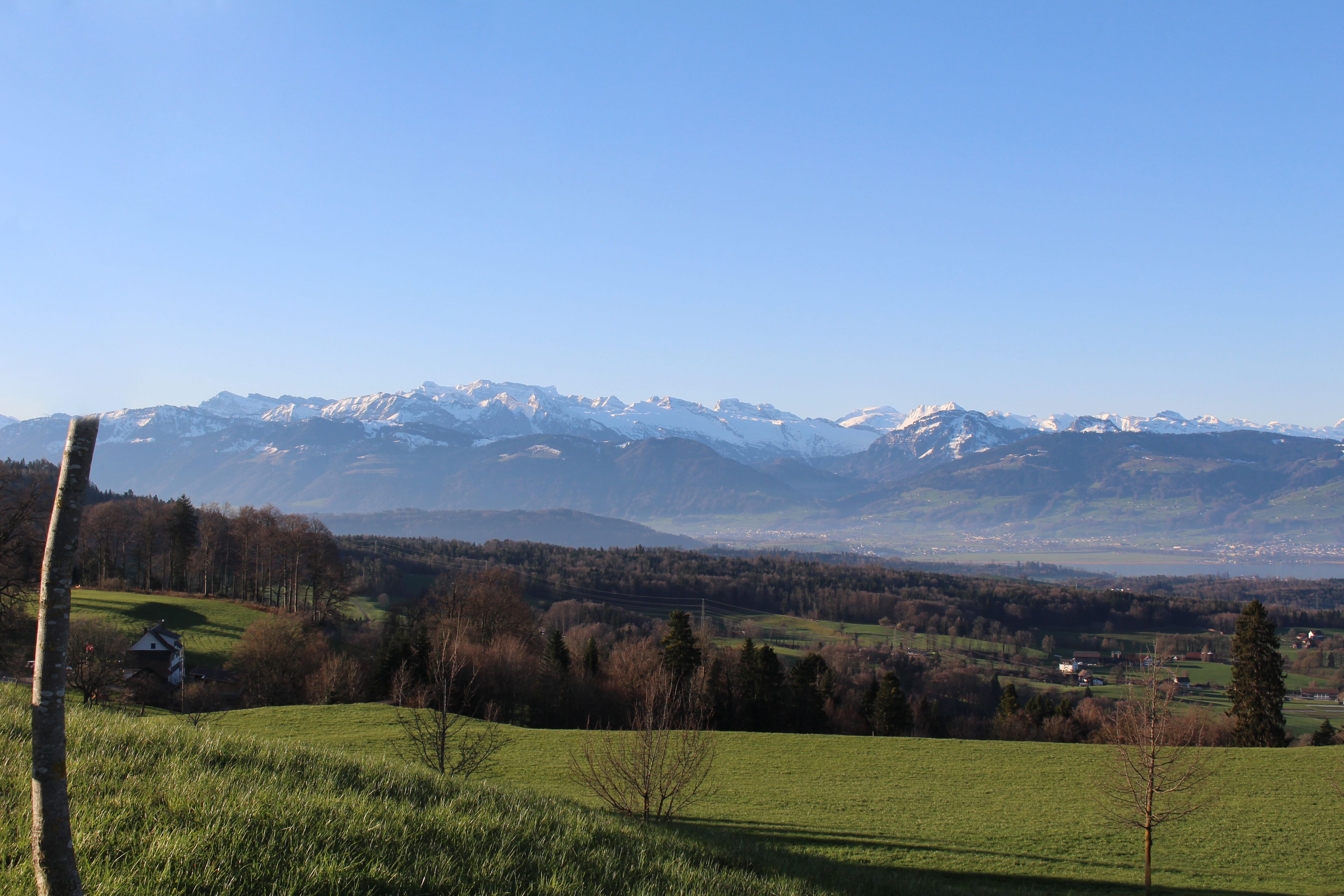 Paisaje rural con una casa en un campo, cielo despejado y montañas nevadas al fondo.